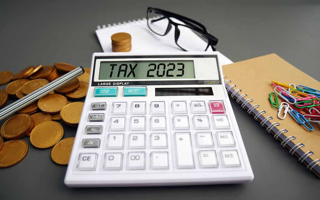 Photo shows a calculator with TAX 2023 displayed on it. In the backgrounds is coins, glasses, paper and paperclips for filing Us Taxes.