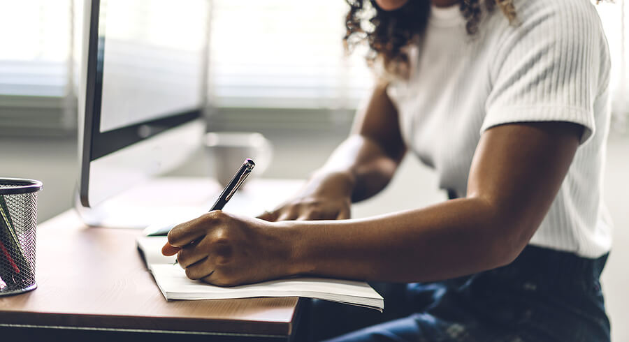 Woman writing in a notebook. She is sitting next to a computer monitor learning about Foreign Earned Income Exclusion, Foreign Tax Credit.