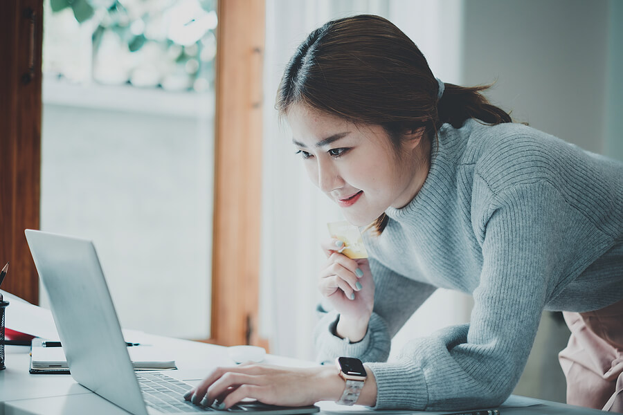 An asian woman leans over her laptop with credit card in hand. She is paying late US expat taxes as an American abroad.