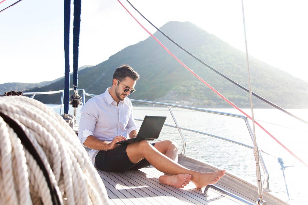 Expat male, sits on a sailboat working at his laptop.